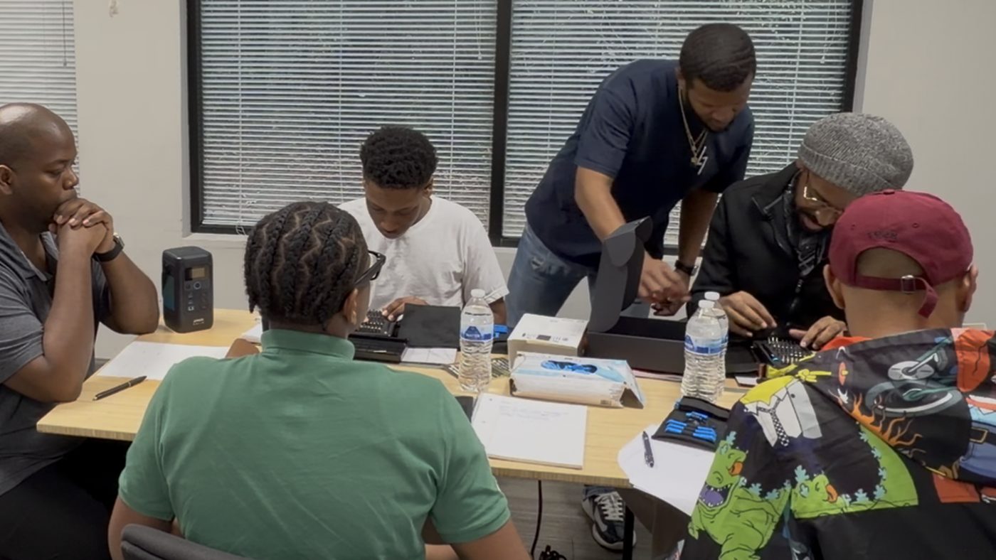 An evening class around the table, the instructor leaning in to help a student with a device