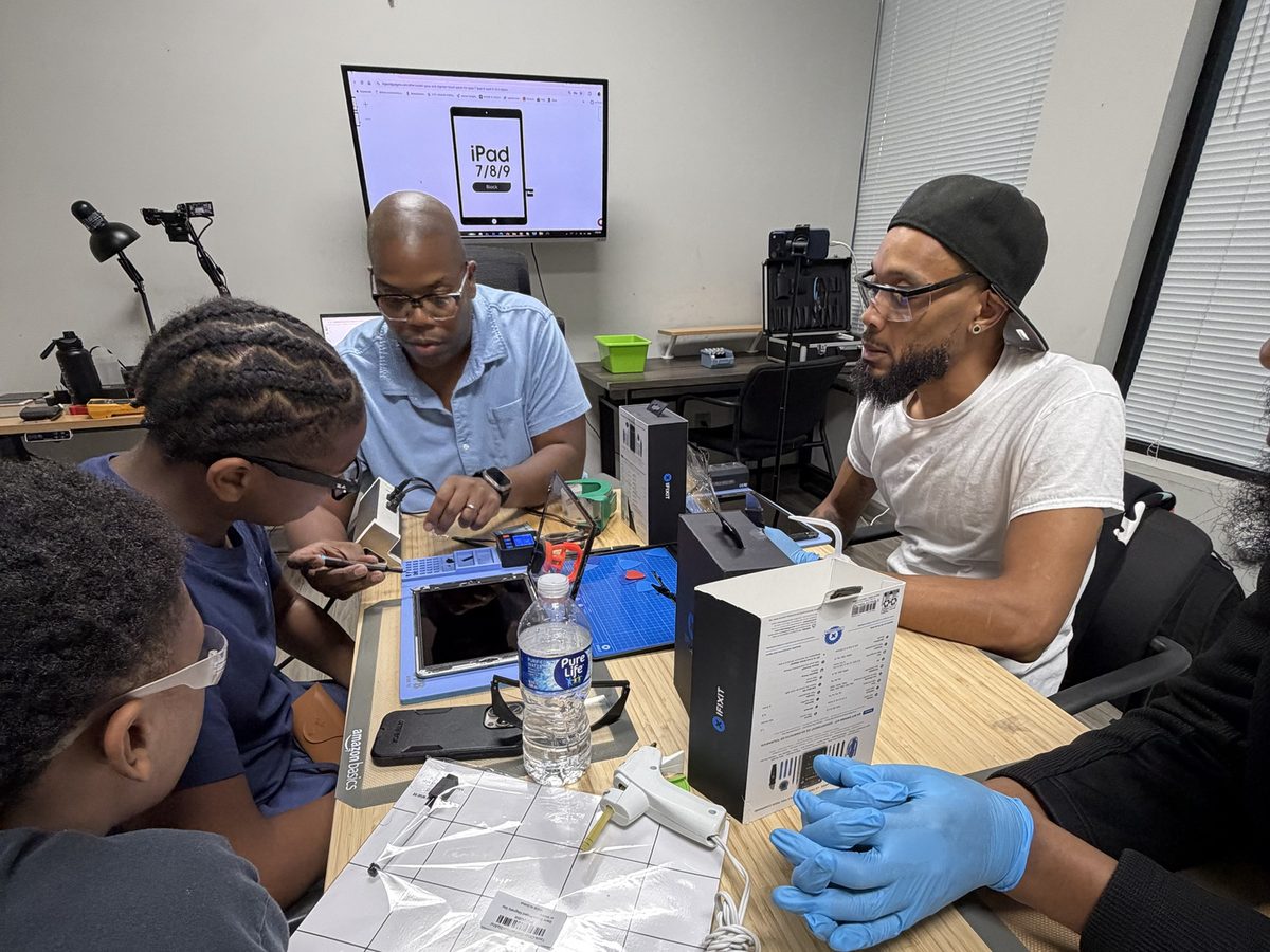 The instructor walking two students through an iPad screen replacement at the bench, the repair guide on the TV behind him