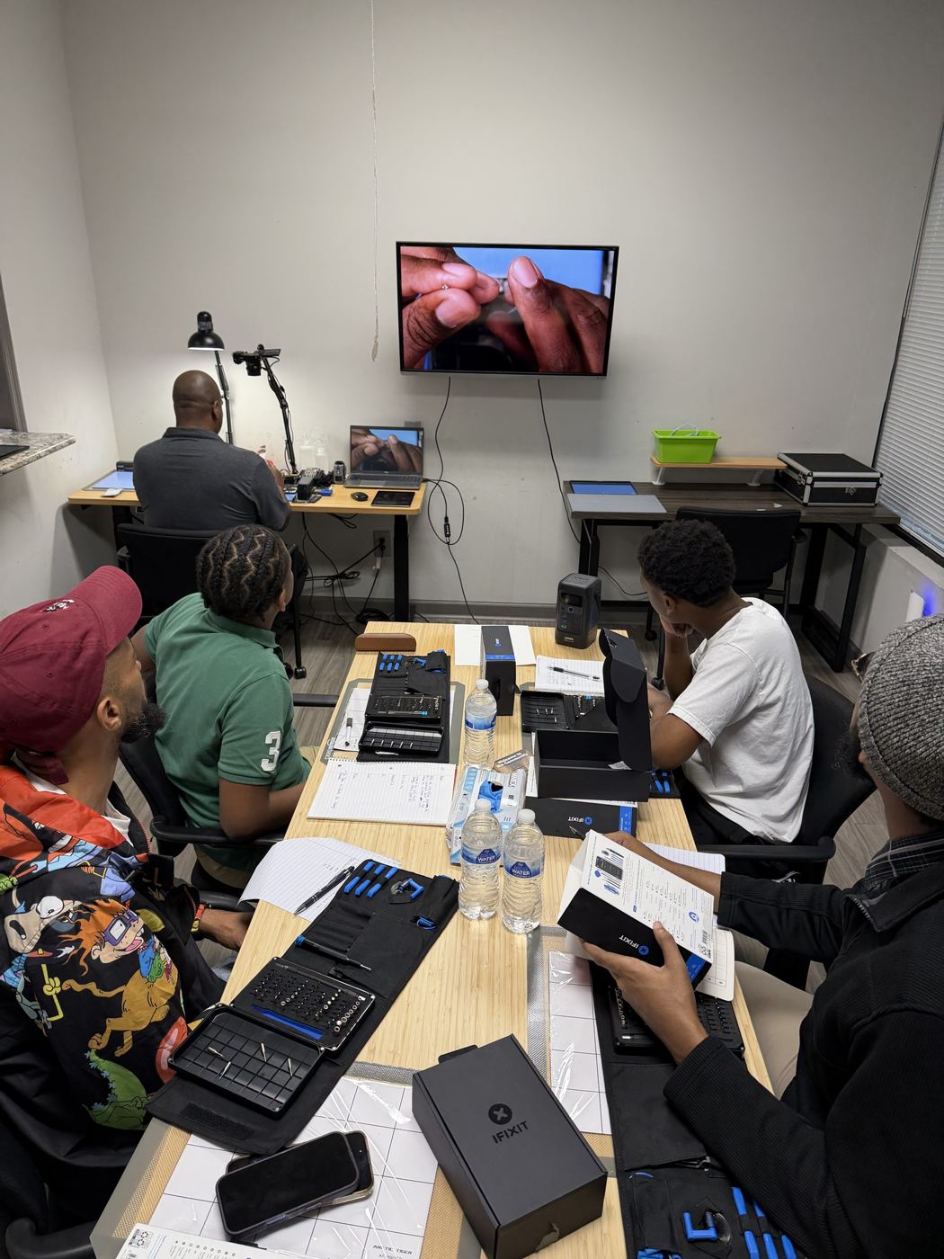 The class watching the bench camera feed on the TV, iFixit kits on the table